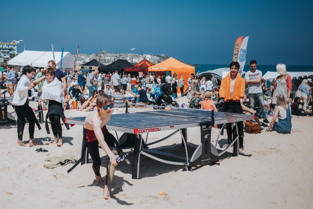 Children play table tennis on a busy beach with food stalls in the background